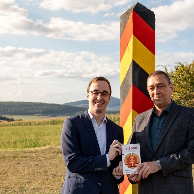 Vorstand Philipp Metzler und Referent Dr. Jens Schöne posieren vor der restaurierten Grenzsäule hinter dem Haus auf der Grenze.